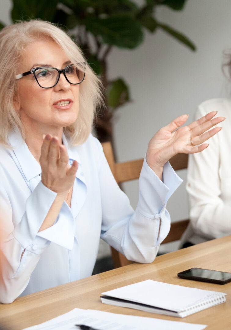 Attractive aged businesswoman, teacher or mentor coach speaking to young people, senior woman in glasses teaching audience at training seminar, female business leader speaker talking at meeting